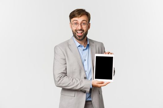 Portrait of happy businessman in gray suit and glasses, introduce application or diagram, showing digital tablet screen and smiling excited, standing over white background.