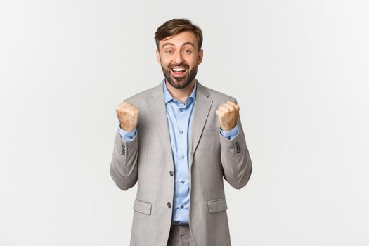 Portrait of relieved and happy businessman with beard, wearing grey suit, achieve goal, clenching fists like a winner, triumphing and rejoicing, standing over white background.