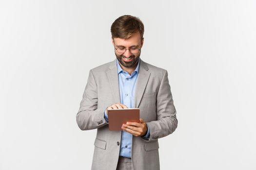 Image of businessman working, looking at digital tablet and smiling pleased, standing in grey suit and glasses over white background.