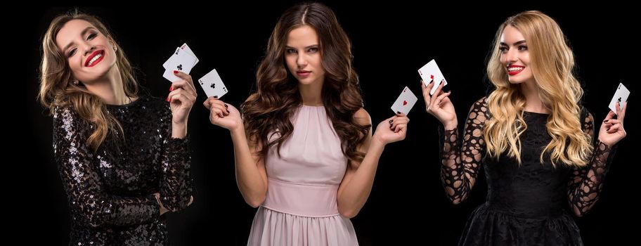 Three beautiful females with bright make-up, in stylish dresses and jewelry. They are smiling, holding aces in their hands, posing on black background. Gambling, poker, casino. Close-up, copy space