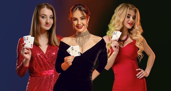 Three gorgeous models with bright make-up, in red and black dresses, stylish jewelry. They smiling, showing aces, posing against colorful background. Gambling, poker, casino. Close-up, copy space