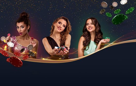 Three gorgeous girls in smart dresses, jewelry. They are holding colorful chips, some of them are flying, posing against blue sparkling background. Copy space for your text or images. Poker, casino