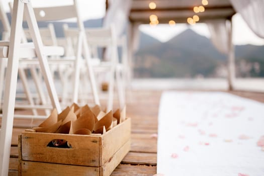 Wooden box with rolled paper bags with flower petals stands on the pier near the chairs. High quality photo