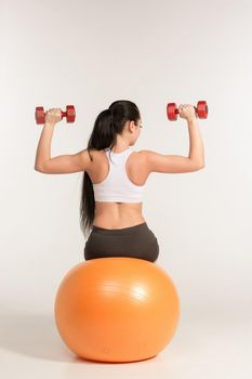 Young beautiful sportswoman doing fitness exercise with dumbbells on pilates ball, at studio shot. Health, beauty and fitness concept.