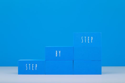 Ladder made of blue toy blocks with written step by step text on white table against blue background with copy space. Concept of business or personal development, success or achieving goals