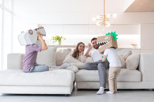Happy family of parents and two children playing dinosaurs at home, children wearing handmade costume mask of cardboard