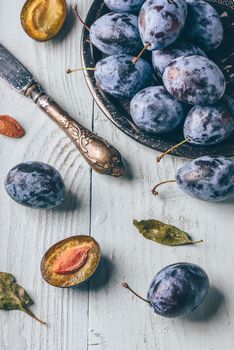 Ripe plums with sliced fruits, leaves and vintage knife over light wooden surface. View from above