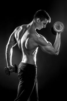 Young handsome man with naked torso and dumbbells in hand on black background. Black and white