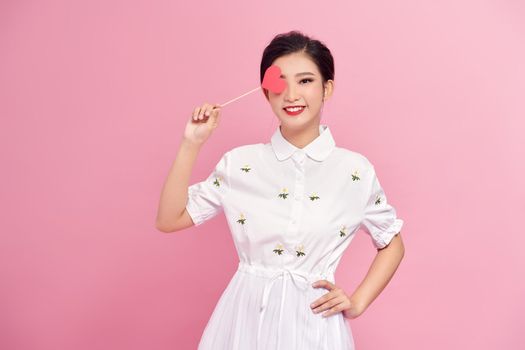 portrait of smiling young woman covering eye with heart on stick isolated