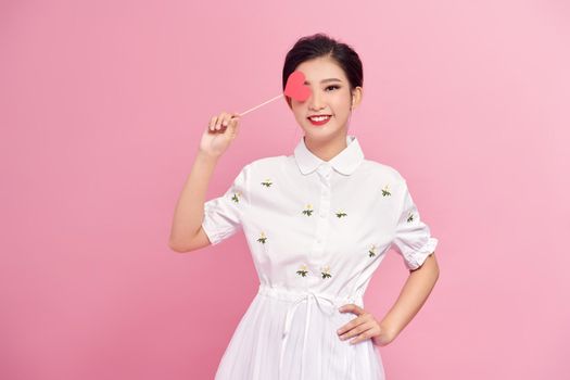 portrait of smiling young woman covering eye with heart on stick isolated 