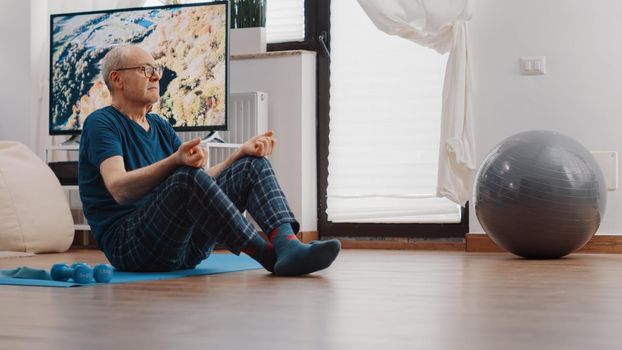 Senior man sitting in lotus position and doing meditation on yoga mat. Zen pensioner breathing to meditate and relax after workout exercise. Old person meditating for wellness and recreation