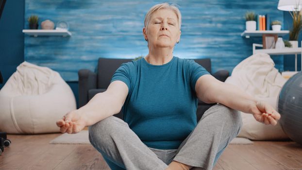 Aged woman doing meditation sitting in lotus position on floor mat. Calm person meditating in recreation before doing physical exercise and gymnastics workout. Elder adult doing yoga pose