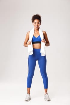 Full length of smiling healthy fitness woman in blue uniform, holding towel and looking satisfied after good workout, standing over white background.