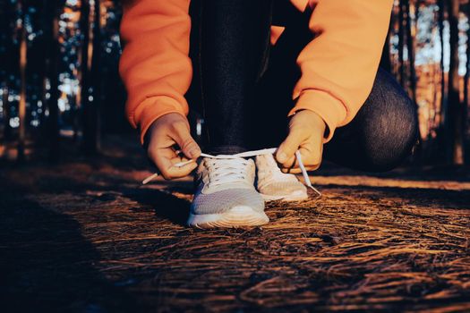 Female runner kneeling and tying shoelace preparing for a run on a forest trail in the morning.