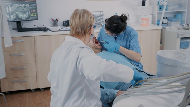 Team of specialists doing drill procedure on patient with toothache, using dental instruments in oral care office. Medic and assistant doing caries inspection, treating man in pain.