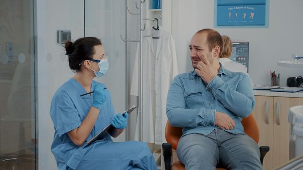 Dentistry nurse taking notes and talking to patient about toothache at oral care clinic. Woman assistant consulting man in pain with denture problems, discussing about dental procedure.