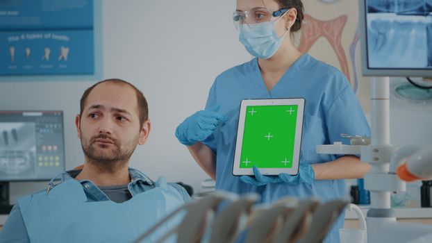 Stomatology nurse vertically holding tablet with green screen, explaining isolated copy space with chroma key background and mock up template to patient at oral care examination.