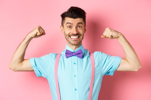 Smiling caucasian man with bow-tie and suspenders, showing muscles and feeling strong, flexing biceps to show-off, standing over pink background.