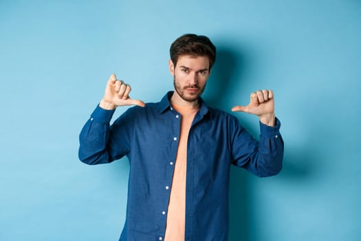 Handsome and confident man with beard, looking at camera and pointing at himself, self-promoting, standing on blue background.