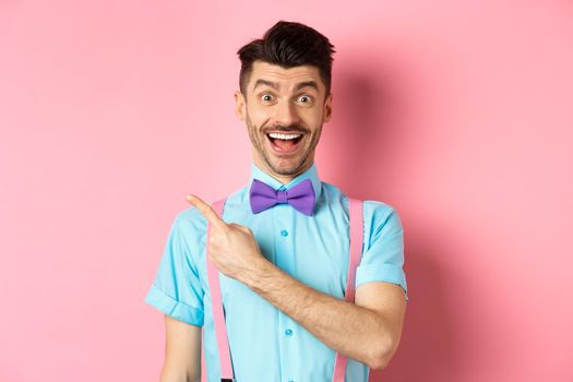 Happy young man checking out promo offer, smiling amazed and pointing finger left at copy space, standing near logo on pink background.