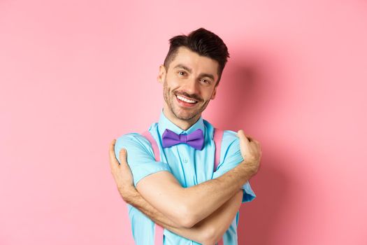 Cheerful young man smiling and hugging himself, wrap hands around own body and looking happy, standing over pink background.