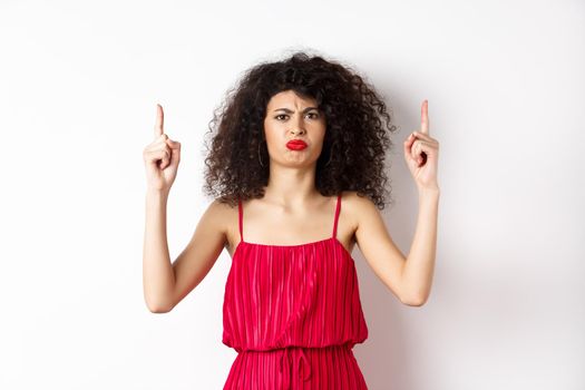 Disappointed frowning woman with makeup, wearing red dress on valentines, pointing fingers up and complaining, standing on white background.