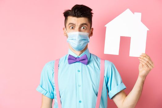 Covid, pandemic and real estate concept. Confused guy in medical mask searching property, showing house cutout and stare at camera worried, standing over pink background.