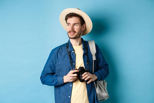 Handsome positive guy in summer hat, holding photo camera and looking aside, tourist taking pictures on vacation, standing with backpack on blue background.