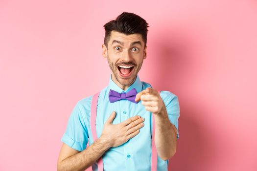 Funny guy laughing at something hilarious, smiling amazed and pointing finger at camera, standing on pink background.