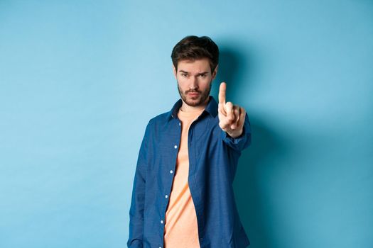Serious young man extend one finger to stop or warn you, prohibit something or disapprove, standing on blue background. Copy space