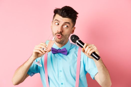 Party and festive events concept. Funny guy singing karaoke, performing song with microphone and drinking champagne from glass, standing on pink background.