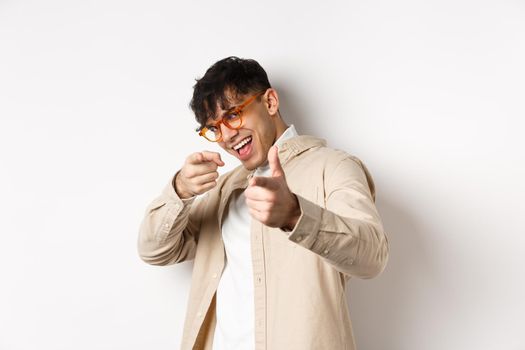 Handsome funny man in glasses smiling, pointing fingers at camera, choosing or inviting you, standing on white background.