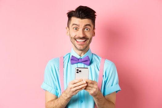 Online shopping. Happy guy in bow-tie, smiling at camera and using smartphone, standing on pink background.