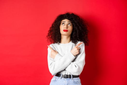 Indecisive female model with red lips and curly hair, looking up troubled with making choice, pointing fingers sideways, choosing between two products, studio background.