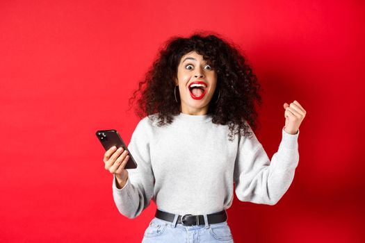 Excited caucasian woman with curly hair, chanting, winning online prize, holding smartphone and triumphing, standing on red background.