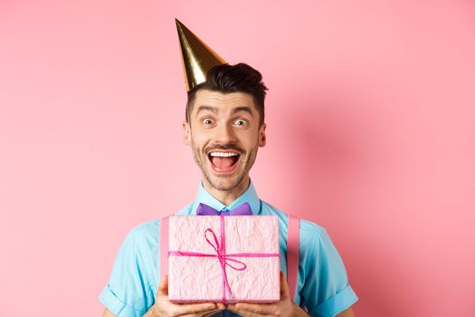 Holidays and celebration concept. Close-up of happy guy in party hat receive birthday gift, scream of joy and cheer, standing over pink background.