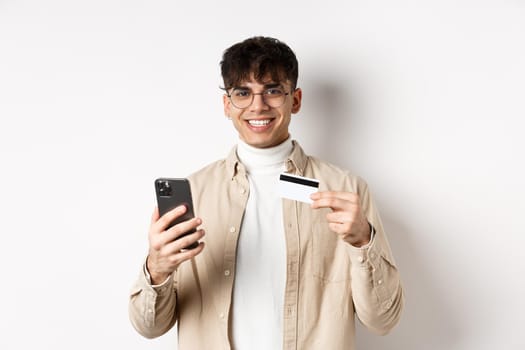Portrait of natural young man in glasses paying in internet, showing smartphone and plastic credit card, standing on white background.