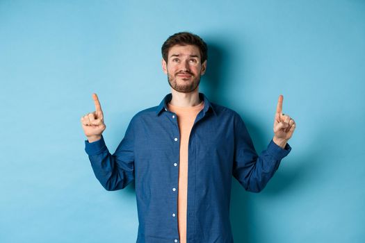 Skeptical young guy smirk and look doubtful, pointing fingers up upset, standing on blue background. Copy space