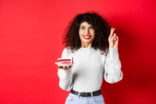 Celebration and holidays concept. Hopeful young woman making birthday wish, holding fingers crossed and looking up, holding b-day cake with candle, red background.