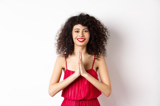 Attractive young woman with red lips, evening dress, saying thank you and smiling, looking grateful with namaste gesture, standing over white background.
