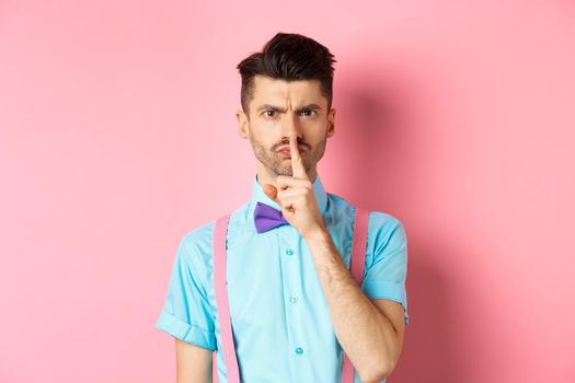 Angry and grumpy man in funny bow-tie shushing at camera, frowning and demand silence, tell to be quiet, scolding someone loud, standing on pink background.