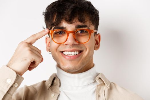 Headshot of smiling happy man showing new eyewear frame, pointing at glasses and looking satisfied, standing on white background.