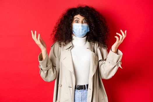 Covid-19, pandemic and quarantine concept. Shocked lady with curly hair and medical mask, raising hands up confused, standing on red background.