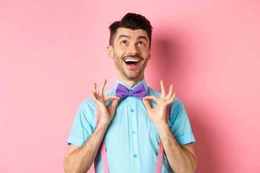 Happy young man with moustache, touching his bow-tie and laughing, looking up at logo, standing on pink background.
