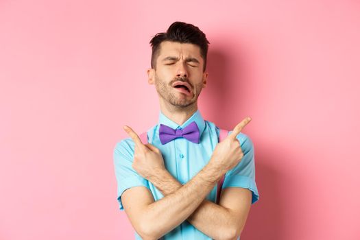 Sad and disappointed young man in bow-tie, crying and pointing fingers sideways, showing two things he wants, sobbing from unrequited love, standing on pink background.