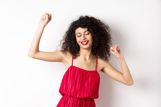 Carefree woman with curly hair, dancing at party, wearing red dress, relaxing with music, standing on white background.