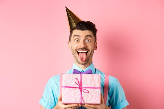 Holidays and celebration concept. Funny guy celebrating birthday, wearing party hat, holding b-day gift and showing tongue with happy face, pink background.