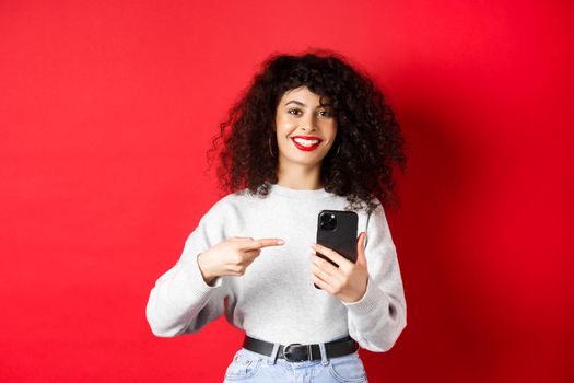 Cheerful pretty lady with smartphone pointing at her phone, smiling satisfied, recommend an app, standing against red background.