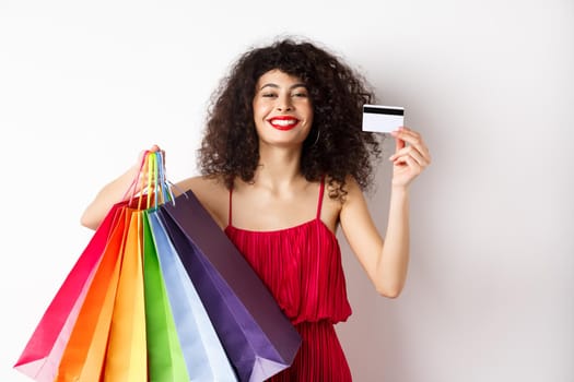 Happy elegant woman in red dress, showing shopping bags and plastic credit card, smiling pleased, standing on white background.