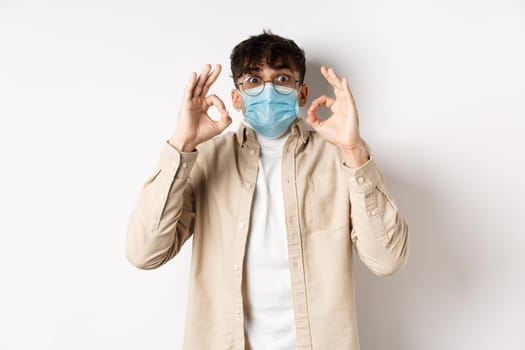 Covid-19, health and real people concept. Excited and impressed guy in sterile face mask showing OK signs in approval, praise cool thing, standing amused on white background.